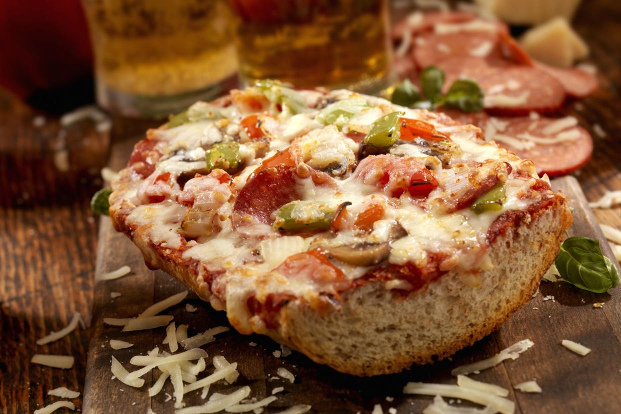 Pizza bite on french bread with pepperoni, mushrooms, red and green peppers with fresh basil and oregano on a wooden cutting board with a couple of beers and ingredients in the background