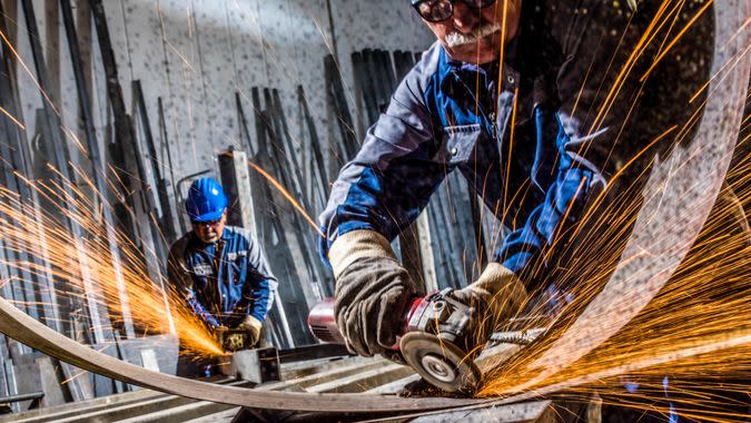 Two senior men using welding torch to cut metal sheet in workshop.