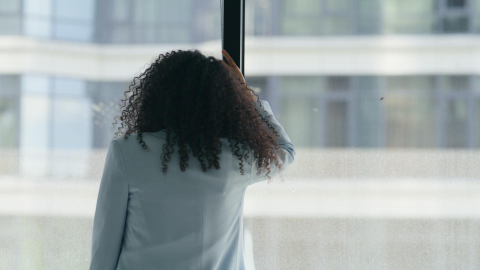 A person with curly hair wearing a formal jacket looks out of a tall window in an urban setting