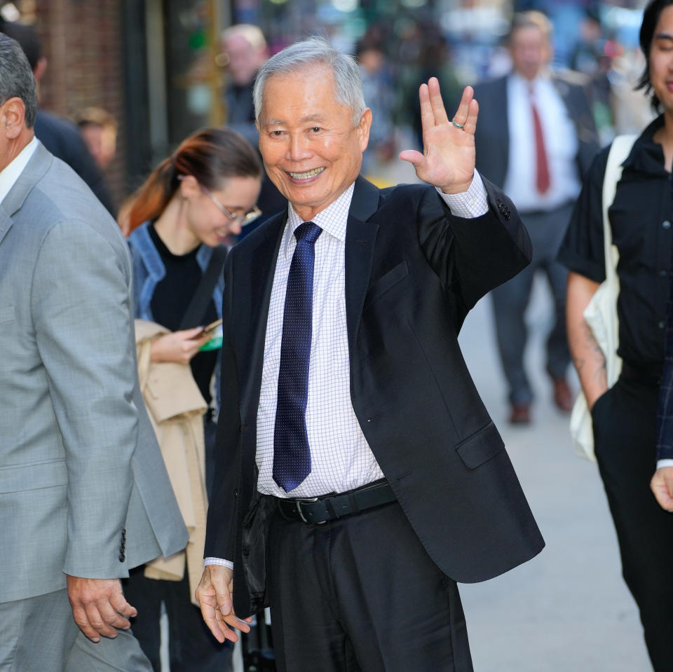 George Takei waves, wearing a suit and patterned tie. Other people, blurred, are around him on the street