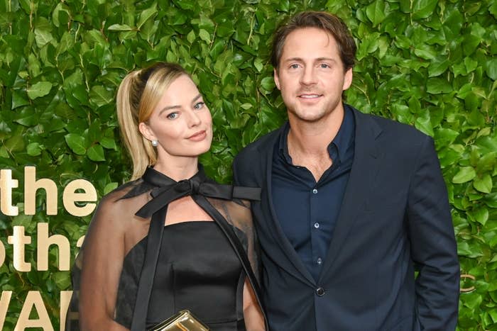 Margot Robbie in a stylish black dress with a sheer overlay and Tom Ackerley in a navy suit pose together in front of a greenery backdrop