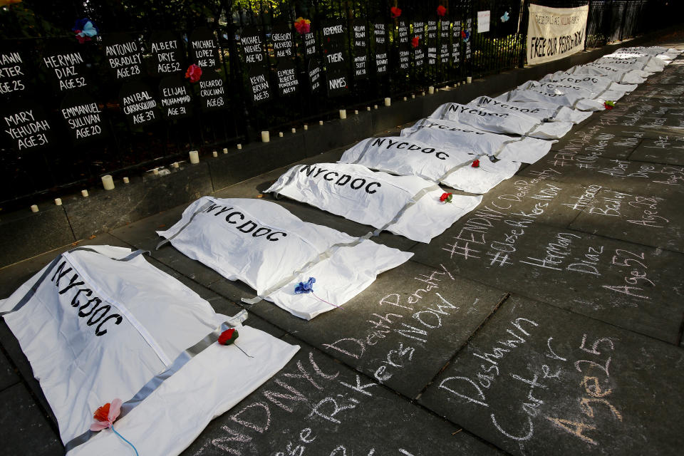 A dozen white body bags marked NYCDOC are laid on the sidewalk. The names and ages of recently deceased inmates are posted behind them and also written in chalk on the sidewalk.