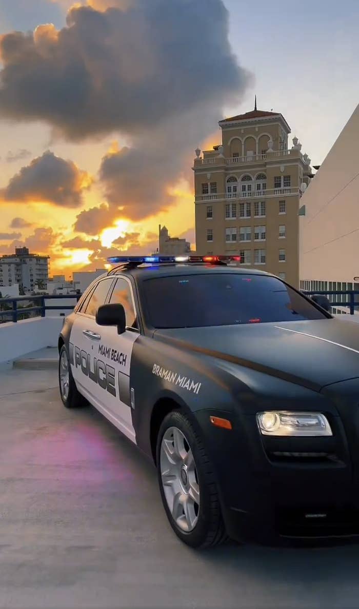 Miami Beach police car with blue and red lights on, parked on a building rooftop at sunset. Background includes a historic building and city skyline