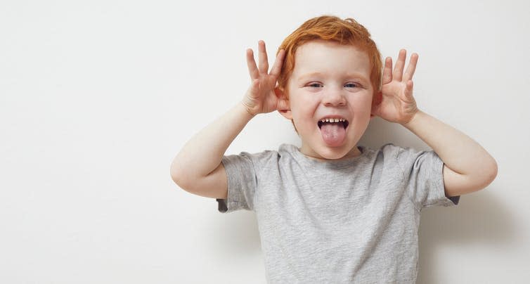 Young boy with his thumbs in his ears, waving hands and sticking his tongue out.