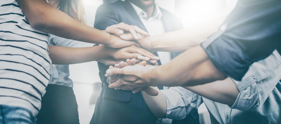 A group of people in a circle stack their hands on top of each other in the middle of the circle suggesting teamwork.