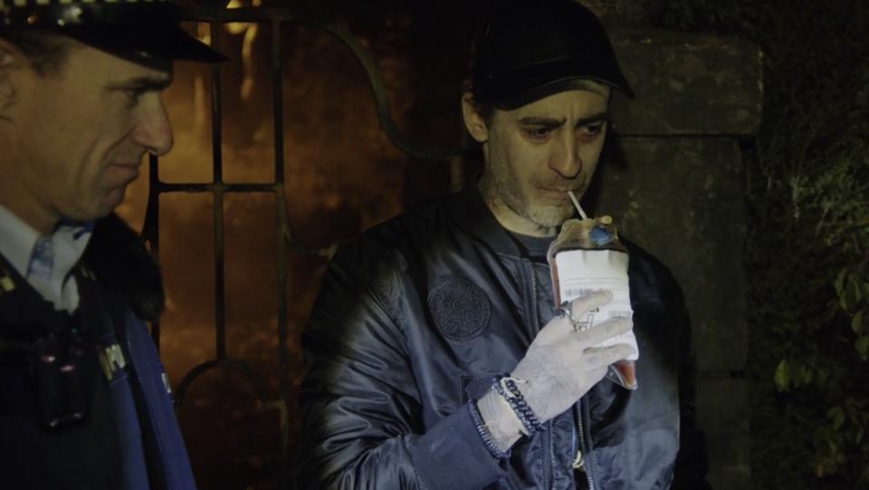 A man drinks blood from a bag with a straw at night while a cop looks on