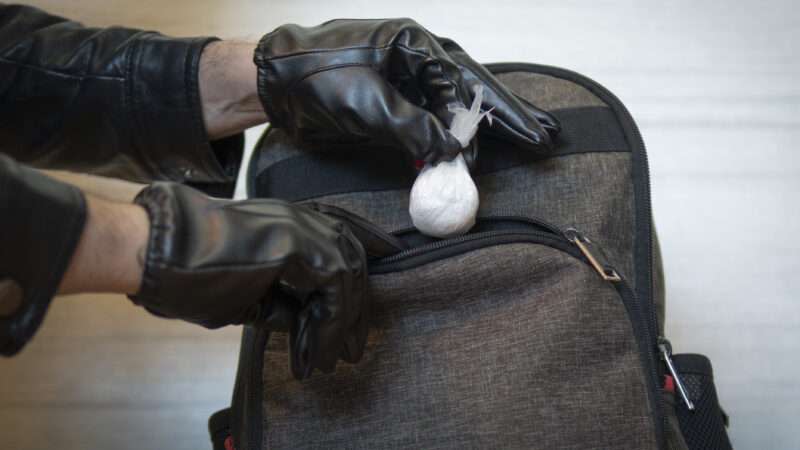 A gloved individual holds a baggy of drugs outside a backpack, as if they are either planting or removing the baggy.