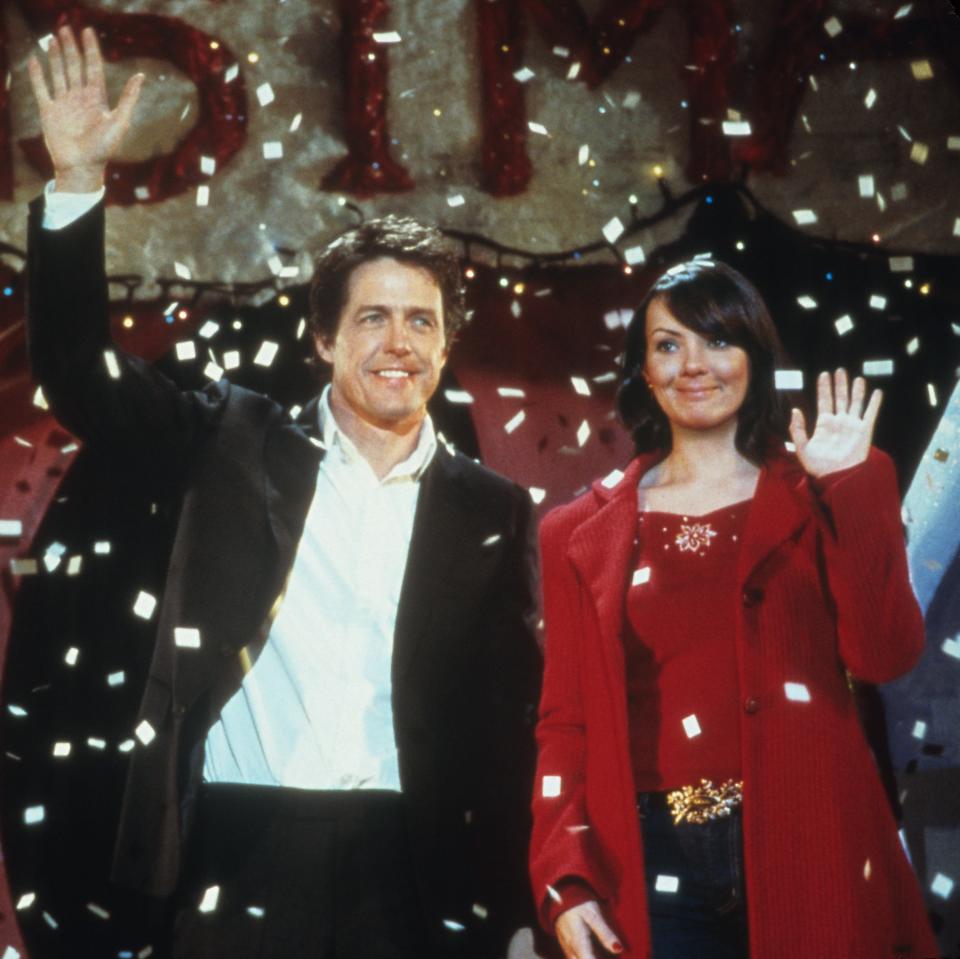 Hugh Grant and Martine McCutcheon wave, surrounded by falling snow-like confetti, in a holiday-themed scene. Hugh wears a black suit, Martine a red coat