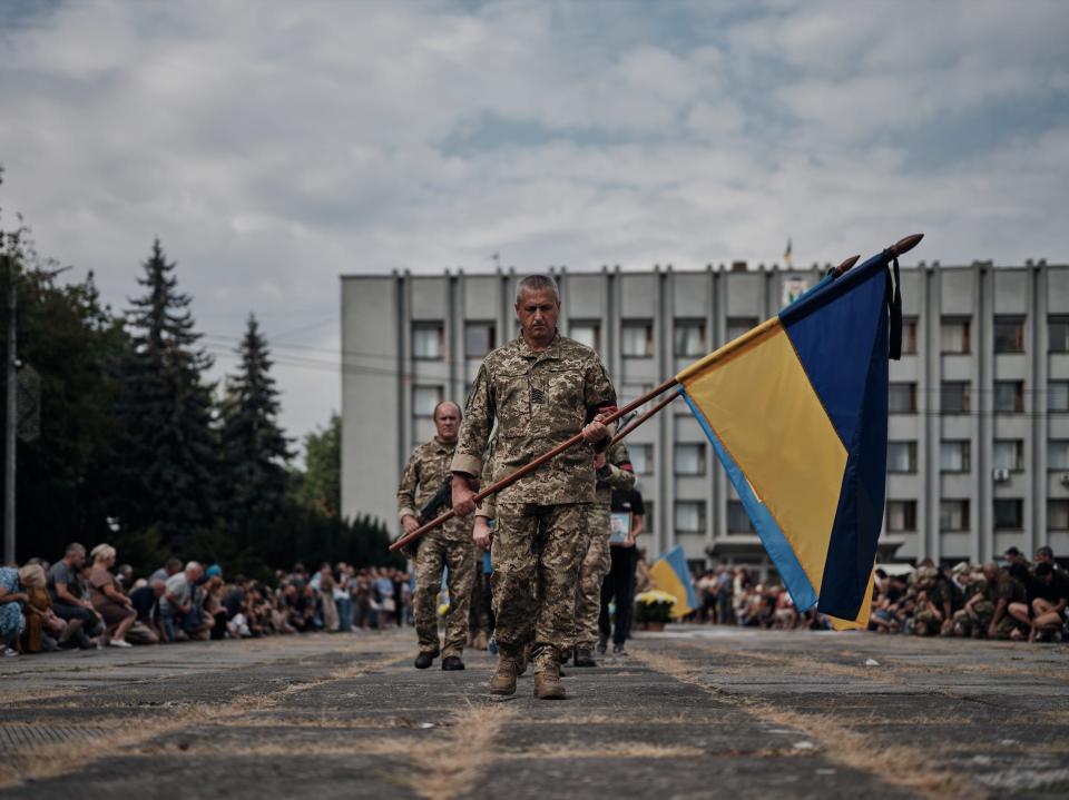 A service member carries a Ukrainian flag at a memorial event for a downed F-16 pilot as hundreds kneel nearby.