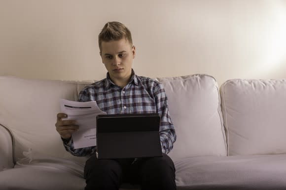 Young male at laptop holding a document.