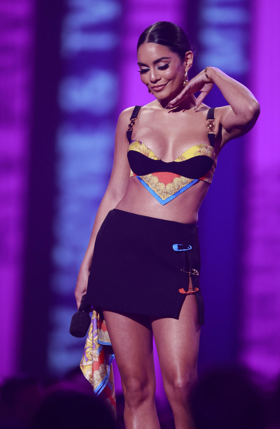 Vanessa Hudgens on stage in a stylish, colorful bralette and black mini skirt, holding a microphone