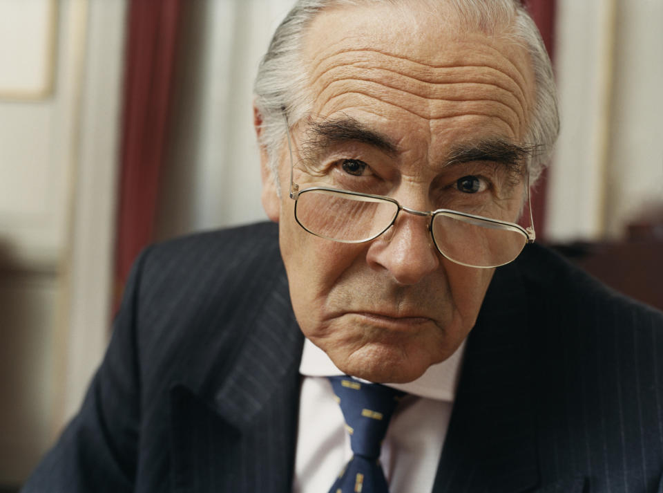 A visibly irritated senior man in a suit with a scowl on his face.