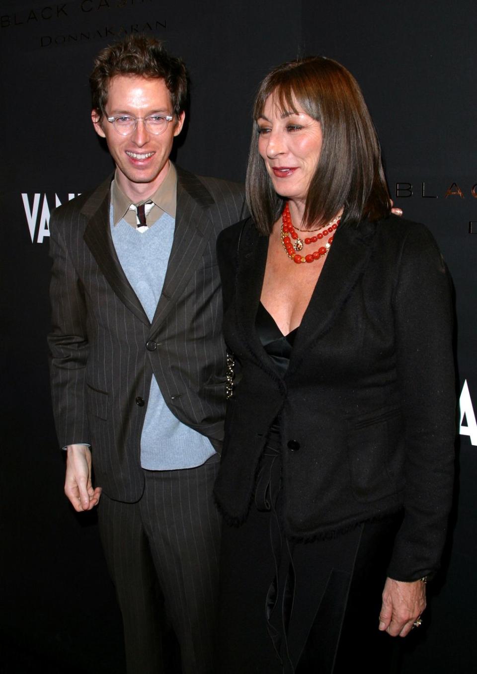 wes anderson, wearing a black striped dress jacket and blue short, puts his hand on the shoulder of angelica jouston, who wears a black pantsuit