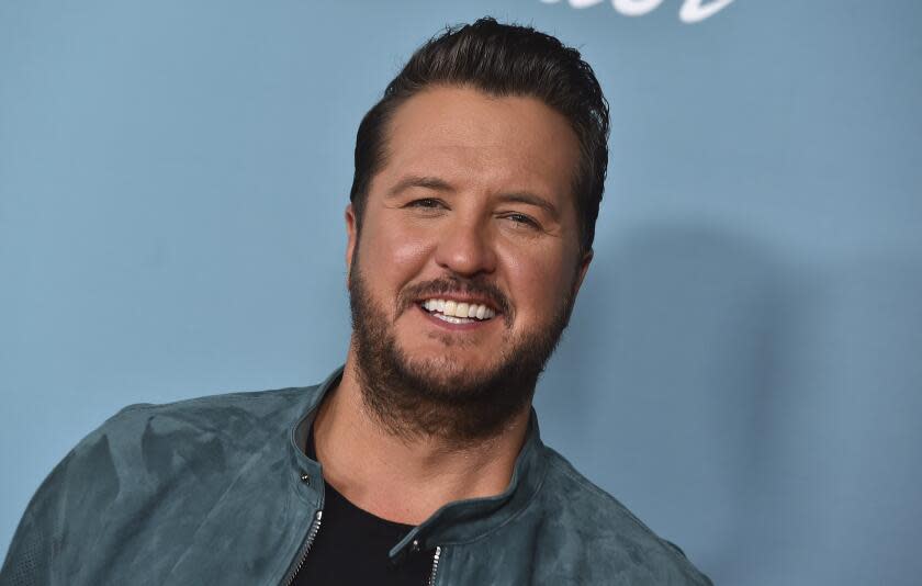 Luke Bryan smiles against a light blue background while wearing a light blue moto jacket