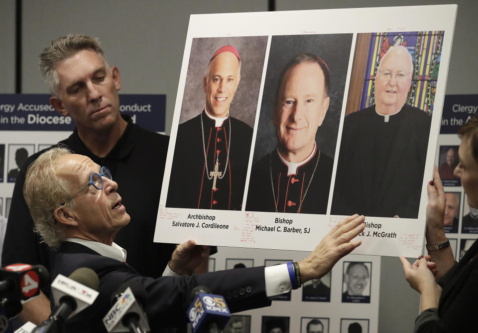 Attorney Jeff Anderson, bottom, and Tom Emens looks toward photos of San Francisco Archbishop Salvatore Cordileone, from left, Oakland Bishop Michael Barber and San Jose Bishop Patrick McGrath at a news conference in San Francisco, Tuesday, Oct. 23, 2018. A law firm suing California bishops for the records of priests accused of sexual abuse has compiled a report of clergy in the San Francisco Bay Area it says are accused of misconduct. (AP Photo/Jeff Chiu)