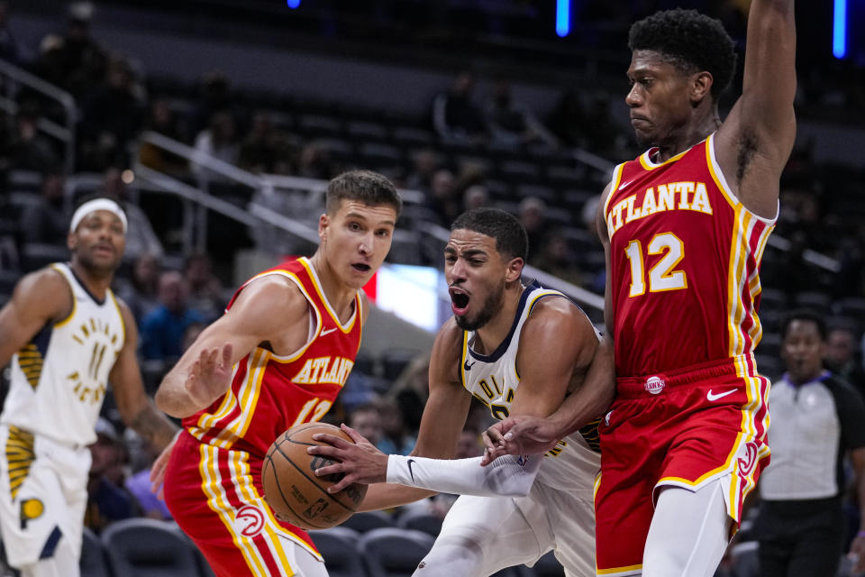 Indiana Pacers guard Tyrese Haliburton (0) is fouled as he cuts between Atlanta Hawks guard Bogdan Bogdanovic (13) and forward De'Andre Hunter (12) during the second half of an NBA preseason basketball game in Indianapolis, Monday, Oct. 16, 2023. Pacers defeated the Hawks 116-112. (AP Photo/Michael Conroy)