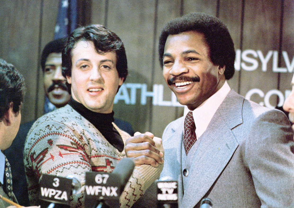 American actors Sylvester Stallone (L) and Carl Weathers grip hands and smile together during a press conference in a still from the film, 'Rocky,' directed by John G. Avildsen, 1976. (Photo by United Artists/Courtesy of Getty Images)