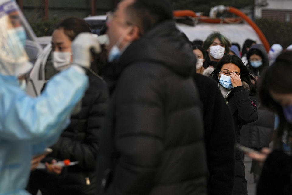 A woman wearing a face mask to protect from the coronavirus looks on as people line up to get a throat swab for the COVID-19 test at a mobile coronavirus testing facility outside a commercial office buildings in Beijing, Monday, Jan. 17, 2022. Beijing's first reported case of the omicron variant has prompted stepped-up measures in the nation's capital, just weeks before it hosts the Winter Olympic Games. (AP Photo/Andy Wong)