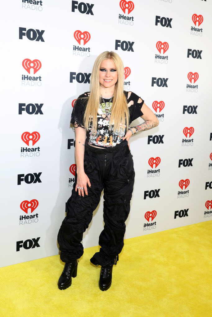 Avril Lavigne standing on a yellow carpet background, wearing a graphic tee, black cargo pants, and boots