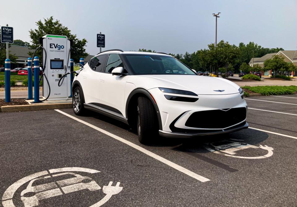 A white Genesis GV60 electric SUV charging at an EVgo station, with trees and sky in the background.