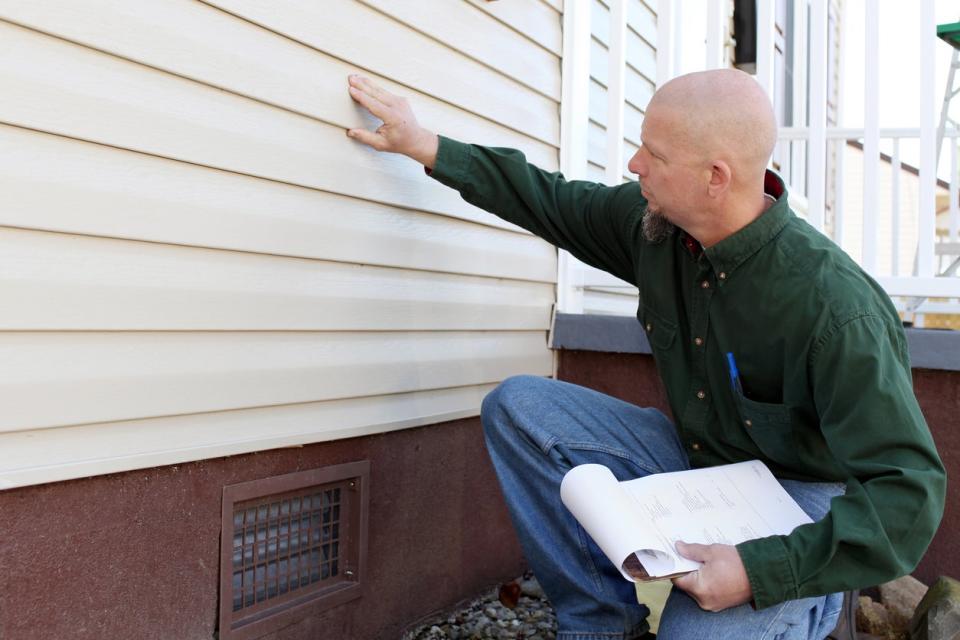 A man holding a clipboard assesses the siding of a home.