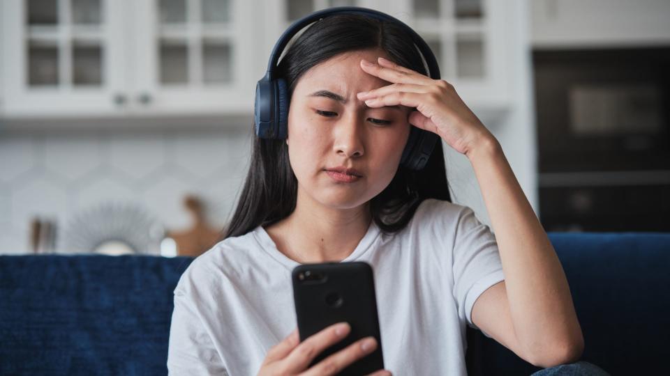 A young woman wearing headphones frowning at her phone