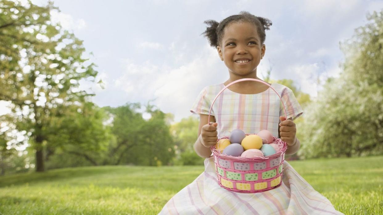 little girl smiling in green field wearing pastel plaid dress with basket full of colorful eggs found during easter egg hunt