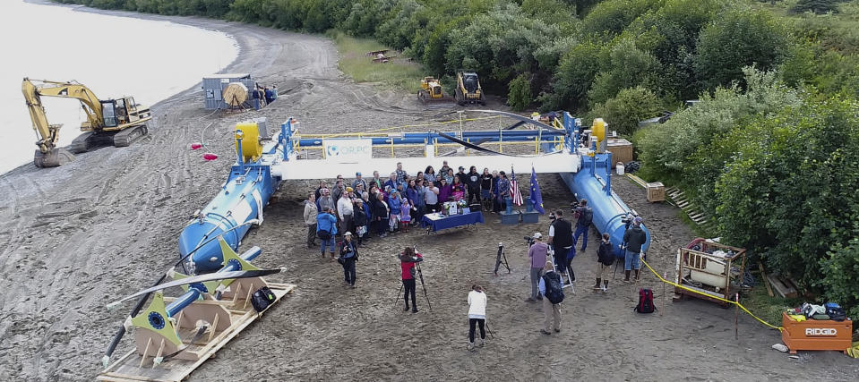 In this Tuesday, July 16, 2019, drone photo released by the University of Alaska Fairbanks and Alaska Center for Energy and Power, Gov. Mike Dunleavy, center rear, poses for photos in front of a Riv-Gen Power System turbine on the bank of the Kvichak River in Igiugig, Alaska. A tiny Alaska Native village is adopting an emerging technology to transform the power of a local river into a renewable energy source. (Amanda Byrd/ University Alaska Fairbanks and Alaska Center for Energy and Power via AP)