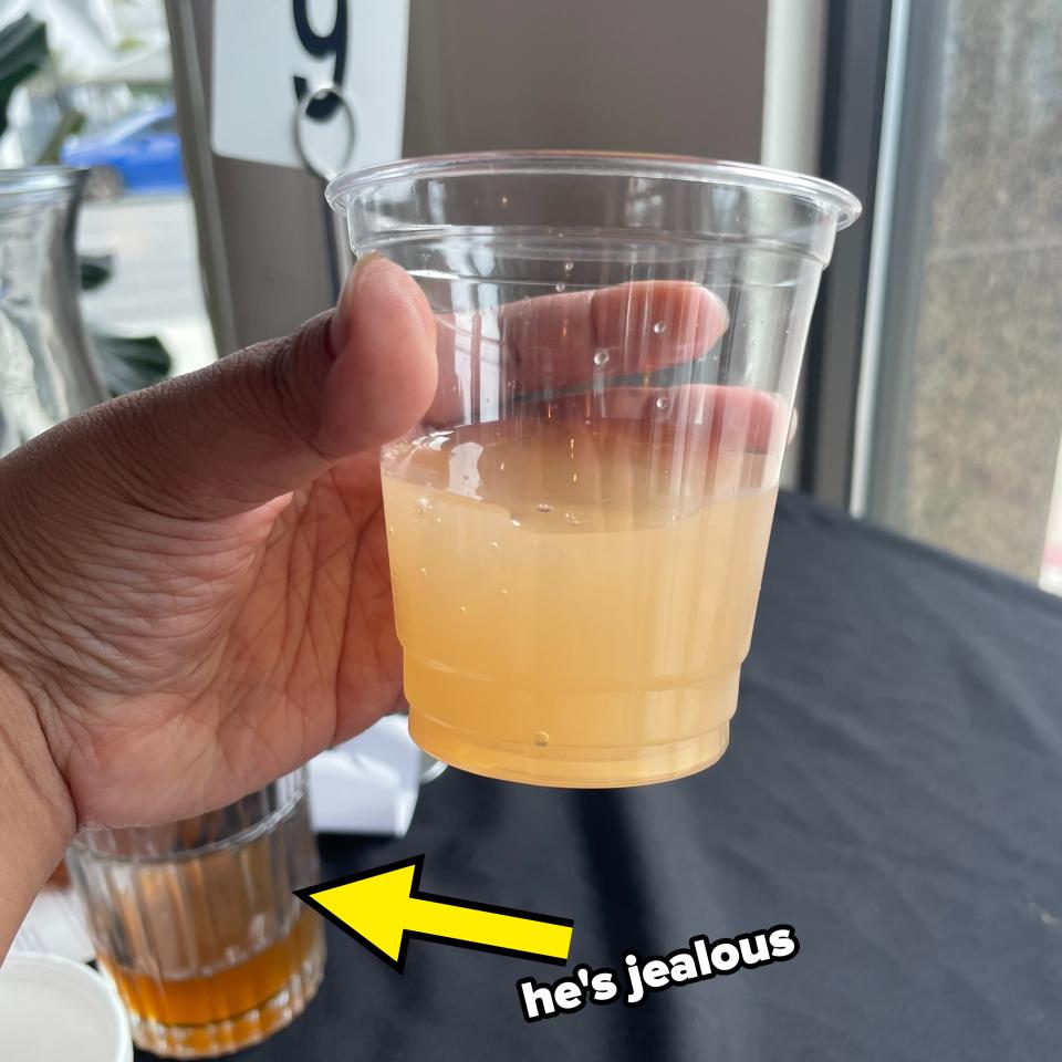 Hand holding a plastic cup of cloudy apple cider in front of a window. Another glass with a similar drink is seen on the table