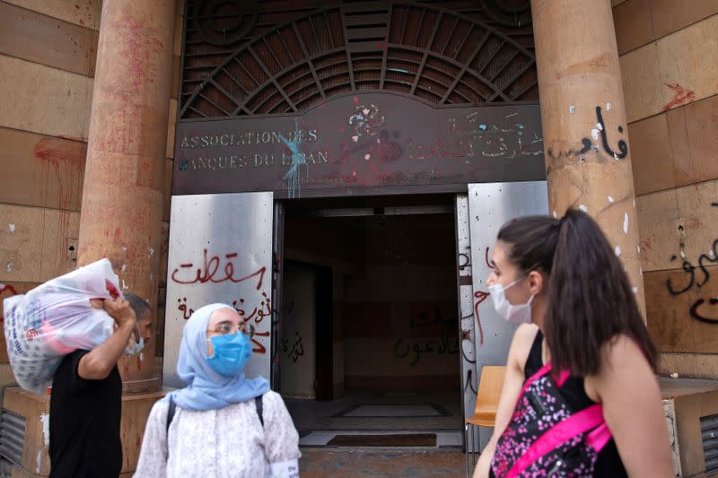 People wearing face masks stand outside the damaged entrance of the Association of Banks in Beirut