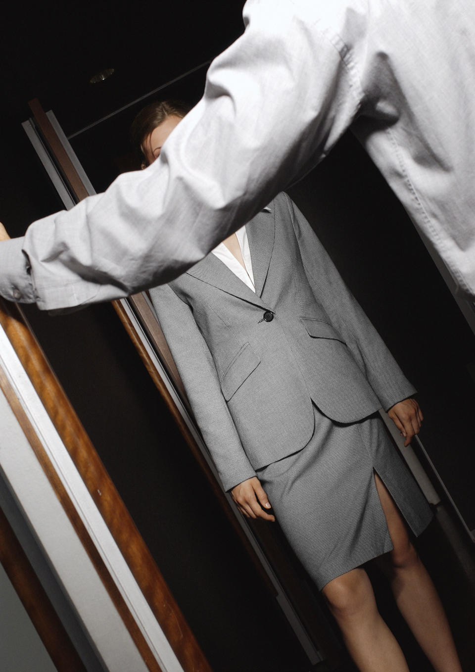 A person wearing a suit opens a door for a woman entering. The woman wears a fitted blazer and knee-length pencil skirt. Their faces are not visible