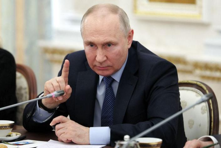 Russian president Vladimir Putin raises a finger in front of a microphone while seated
