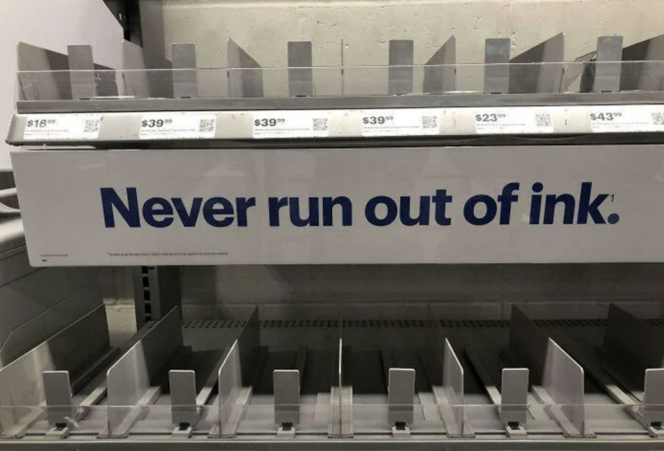 Shelves for printer ink cartridges with an empty stock and a sign stating 'Never run out of ink.'