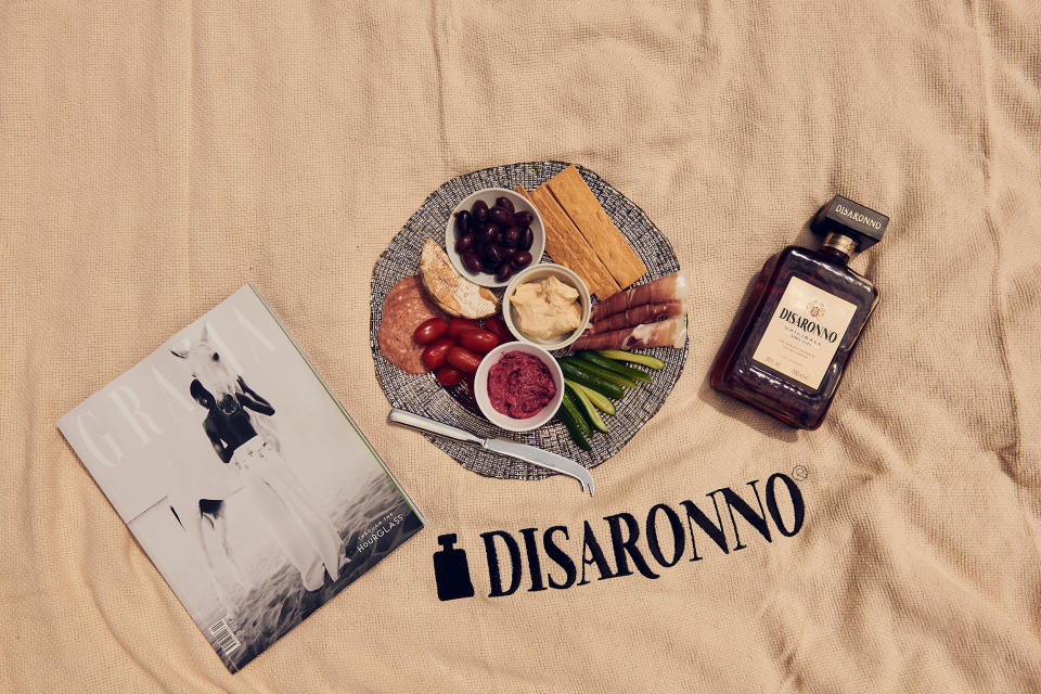 Tela con iscrizione Disaronno stesa con una bottiglia di liquore, un piatto di antipasto e la rivista Grazia