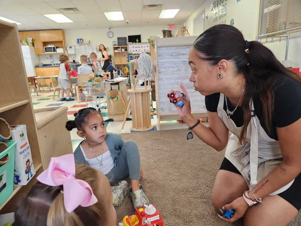 Child care worker Marci Then helps her daughter, Mila, 4, put away toys to get ready for circle time at the Little Learners Academy in Smithfield, R.I. (Elaine S. Povich/Stateline)