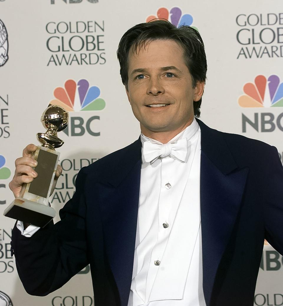 michael j fox holds a golden globe trophy in his right hand and smiles as he looks past the camera, he is wearing a black suit jacket, white shirt and white bow tie and stands in front of a golden globe awards backdrop