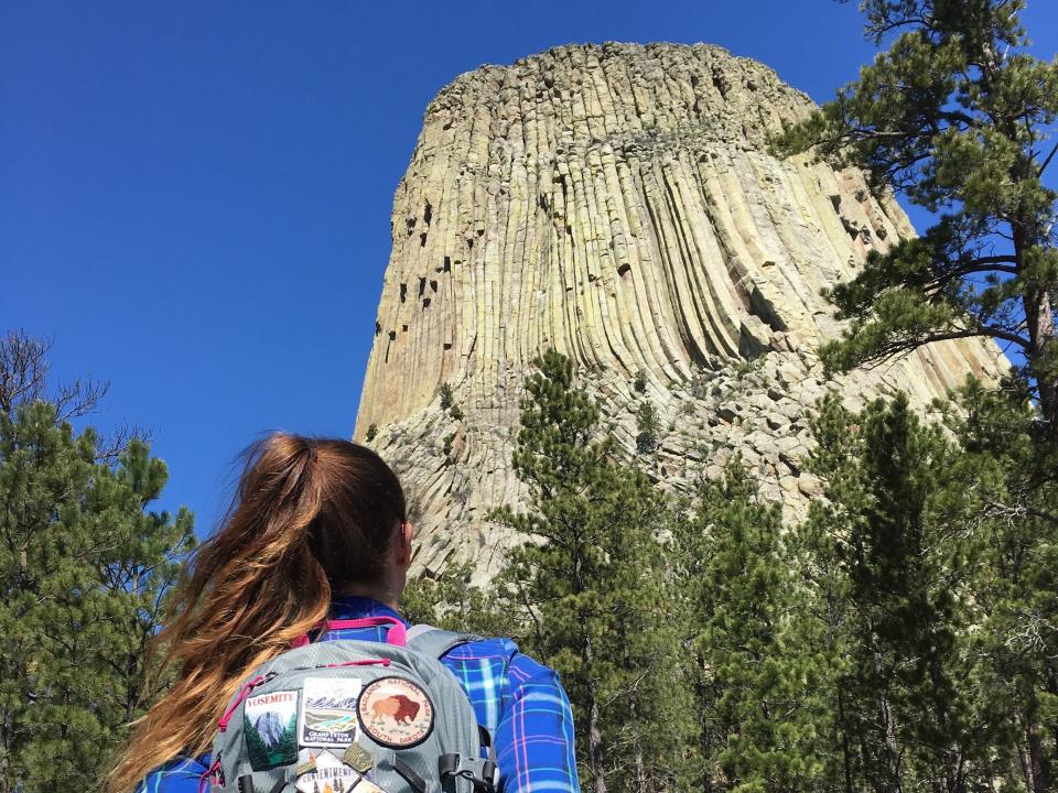 Emily, wearing a flannel and a backpack with patches, looks up at Devils Tower National Monument in Wyoming.