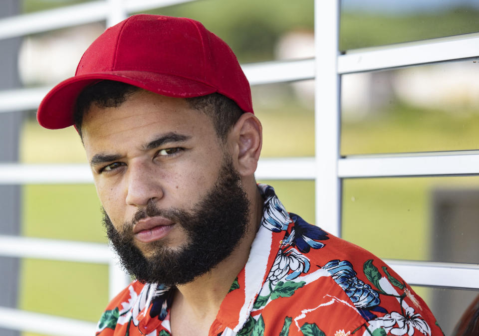 A generic shot of Kurt Henderson (Calvin Demba), wearing a red baseball cap and a loud Hawaiian shirt, standing in front of a grille at the airport