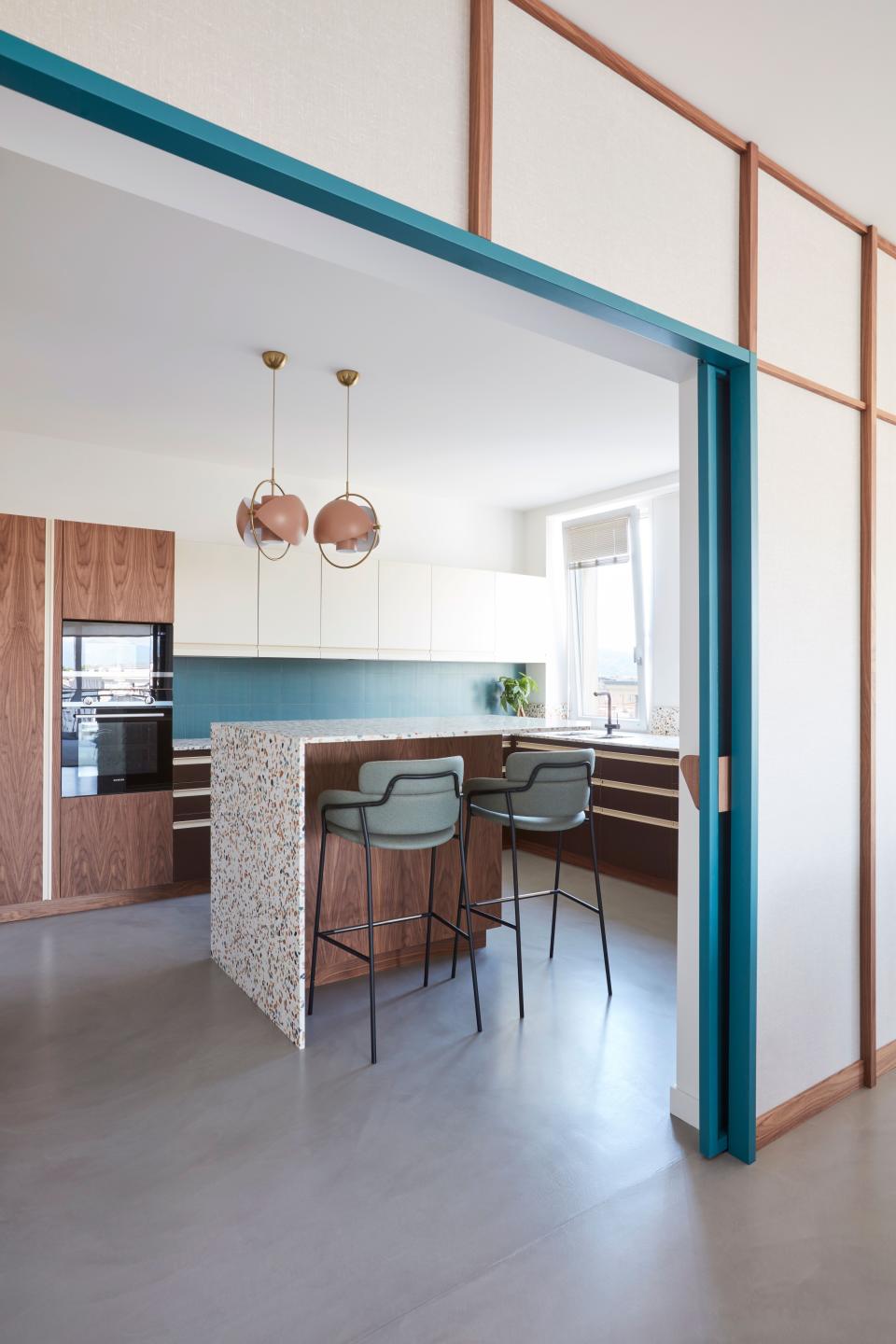 In the kitchen, the use of American walnut helps highlight the blue elements such as the two stools and backsplash. The central island is covered with terrazzo by Agglotech.