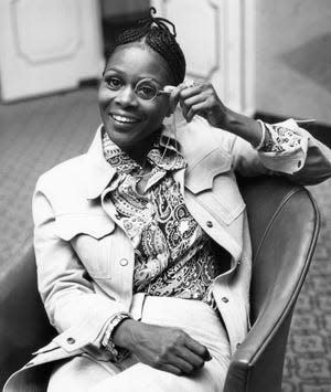 Cicely Tyson poses with a monocle in a chair, sporting cornrow hairstyle, printed shirt and jacket.
