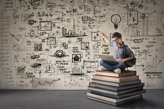 A man sitting on top of a pile of books pointing at a wall covered with diagrams.