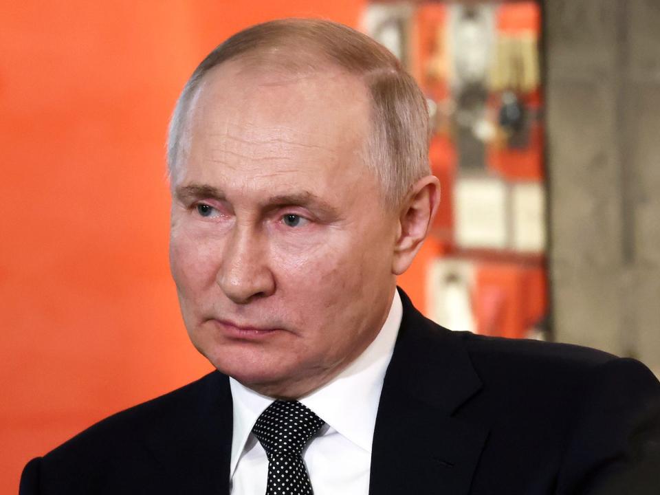 A headshot of Russian President Vladimir Putin standing in front of an orange background.