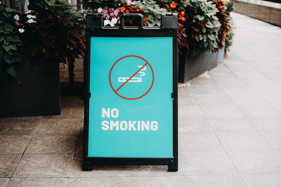 A sidewalk sign with the message "No Smoking" and an icon of a cigarette crossed out, placed next to planters with flowers