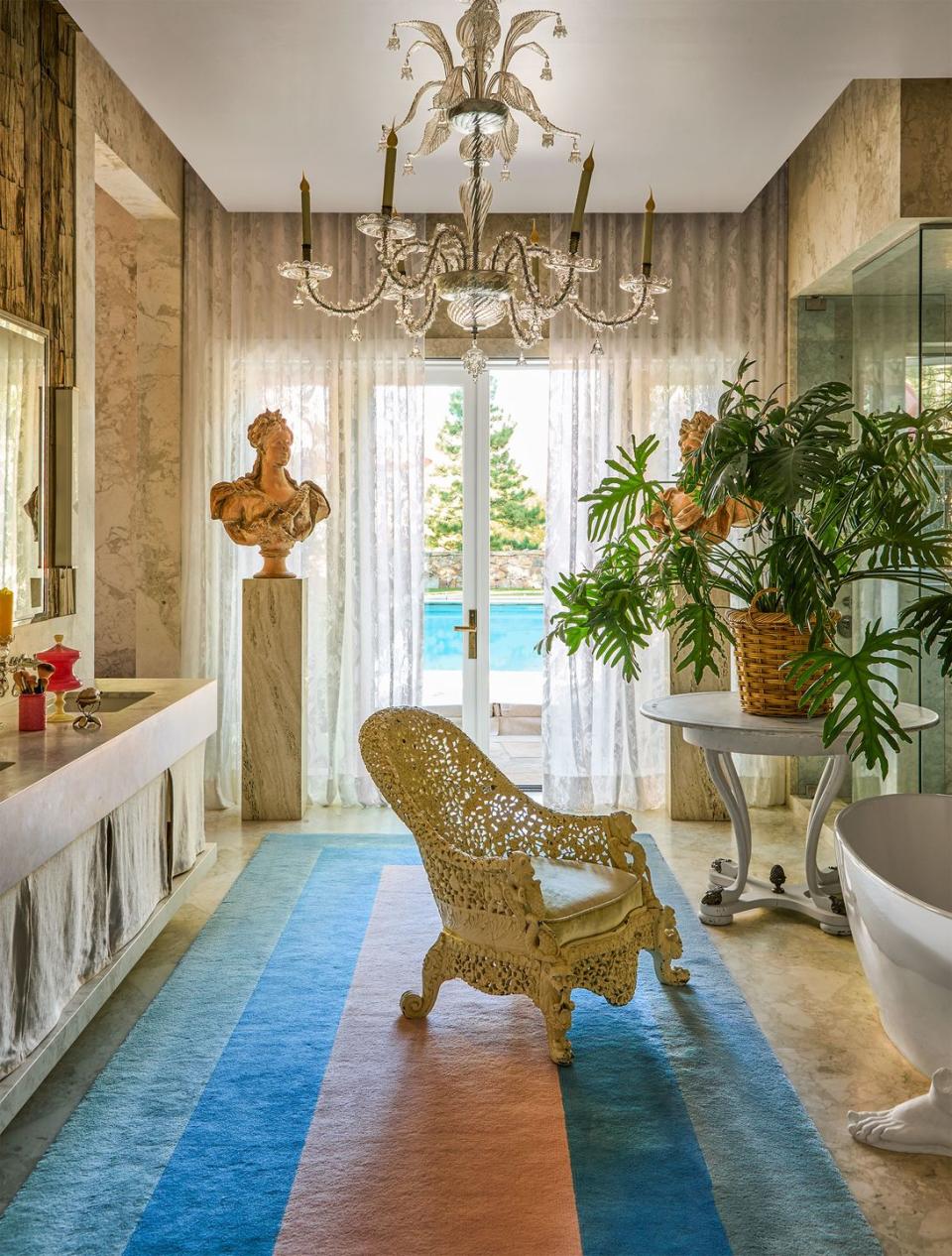 a footed bathtub, an antique chair, counter and twin sinks, plinth with bust sculpture, round table with large plant in basket planter, glass chandelier, transparent curtains, glass doors open to pool