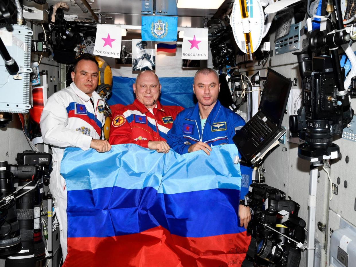 three cosmonauts pose with blue and red striped flag inside space station