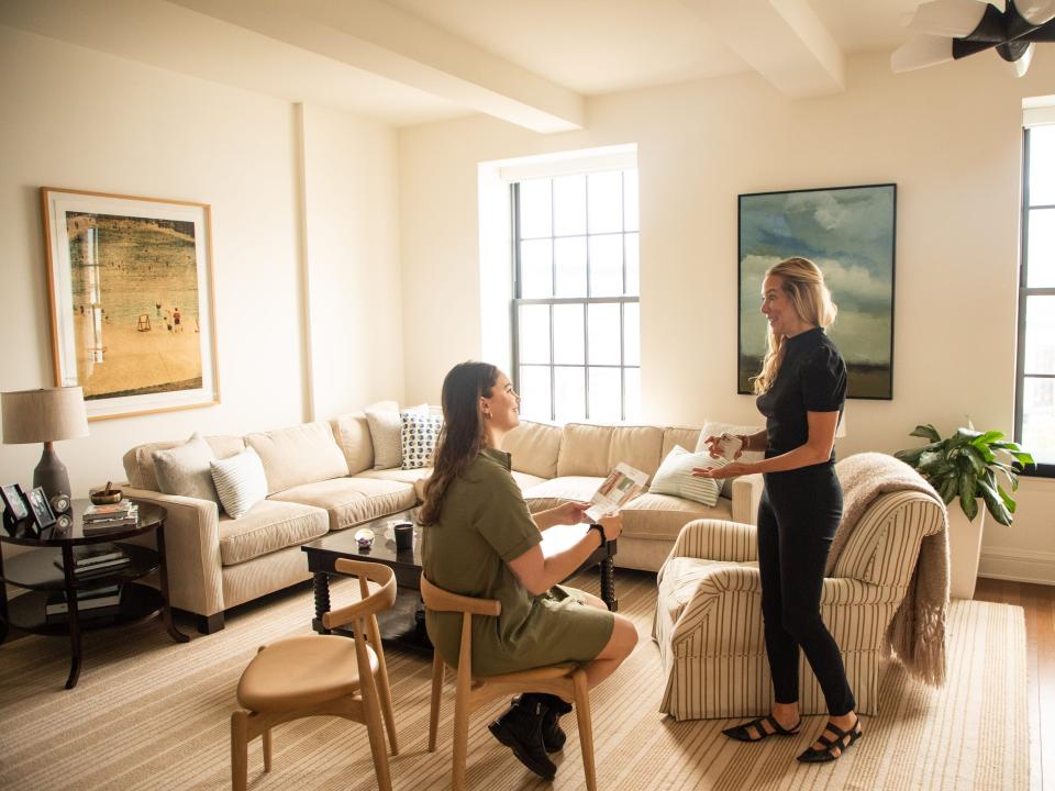 One woman walks towards another woman sitting in a chair in a living room.