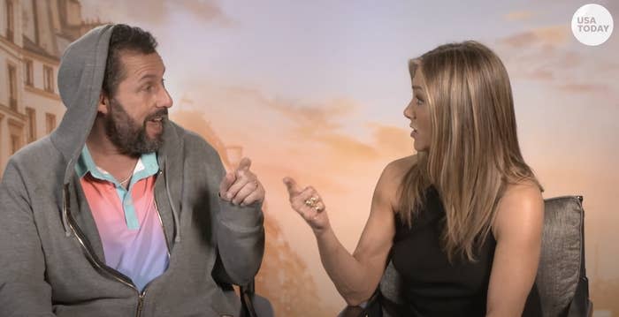 Adam Sandler and Jennifer Aniston sit and gesture toward each other during an interview with USA Today. Sandler wears casual attire, while Aniston is in a stylish gown