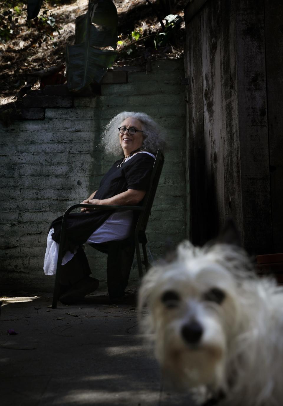 Alison Saar, seated in a chair, smiles as her fuzzy dog Crumb makes an appearance in the foreground of a picture