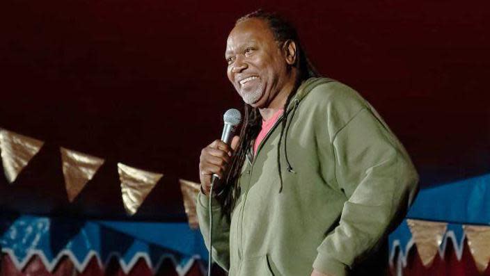 Reginald D Hunter on a stage wearing a green hoody holding a microphone