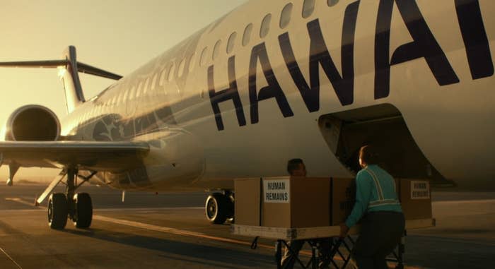 A large box labeled human remains is placed into an airplane's cargo hold by two employees